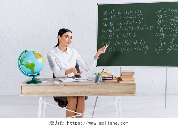 坐在讲台桌上的女人在讲解课题微笑的老师指着地球仪旁边的黑板和教室里的书本 